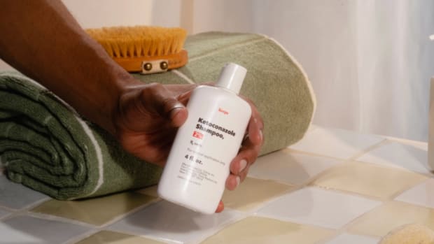 A man in a bathroom holding a bottle of Keeps ketoconazole shampoo for hair growth.